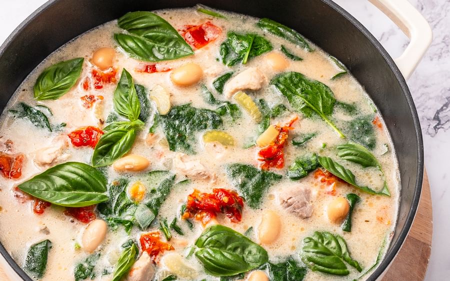 Creamy white bean and spinach soup in a pot with basil, tomatoes, and bread slices nearby.