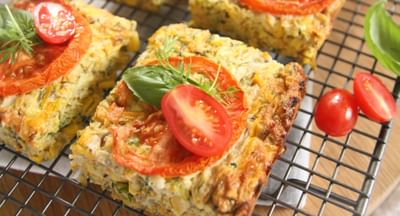 Baked veggie frittata squares on a wire rack, topped with tomato slices and fresh herbs.