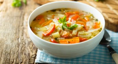 White bowl of vegetable soup with carrots, potatoes and cabbage, garnished with parsley on a napkin.
