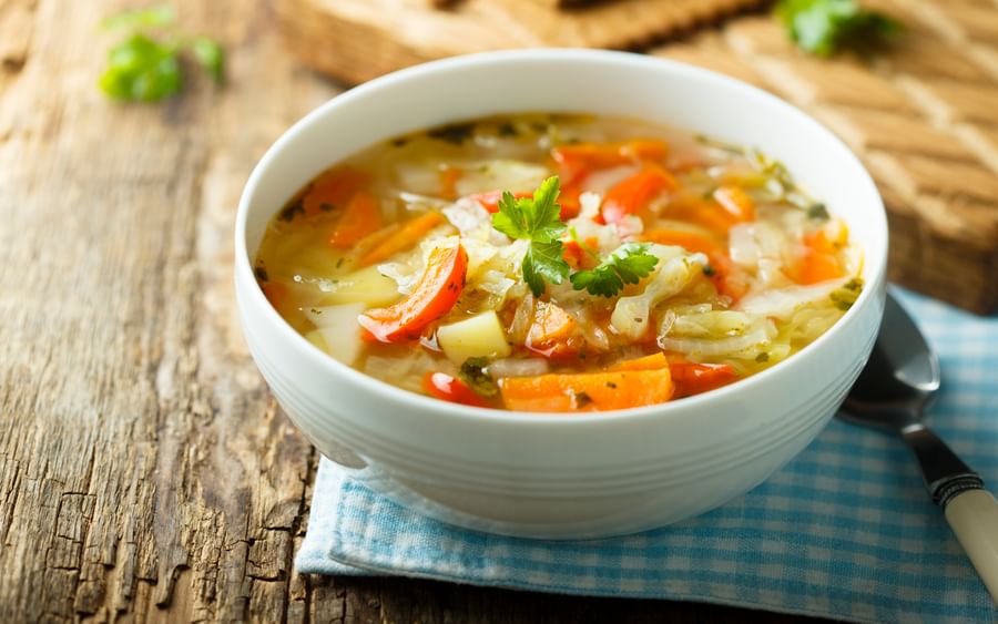 White bowl of vegetable soup with carrots, potatoes and cabbage, garnished with parsley on a napkin.