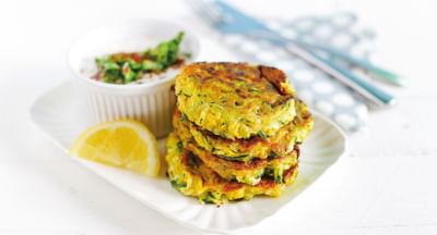 Stack of golden zucchini fritters with herbs, lemon wedge and dip on a white plate.