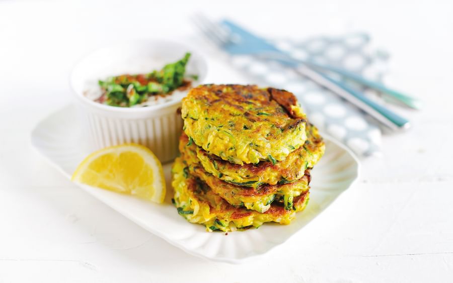 Stack of golden zucchini fritters with herbs, lemon wedge and dip on a white plate.