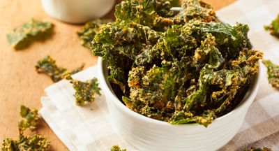 Bowl of crispy kale chips on a light checkered cloth, with scattered kale pieces.