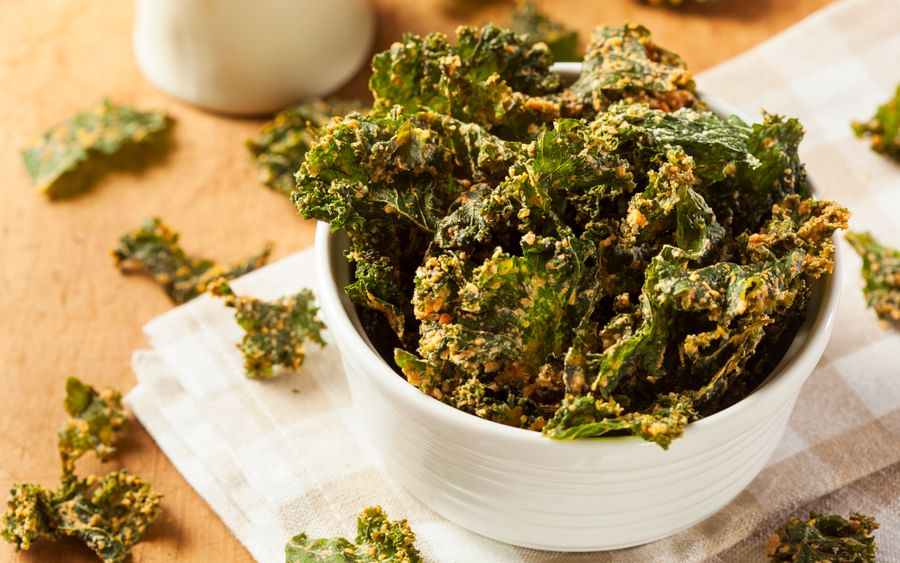 Bowl of crispy kale chips on a light checkered cloth, with scattered kale pieces.