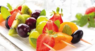 Assorted fresh fruit on skewers on a white platter: strawberries, kiwi, grapes, watermelon, orange.