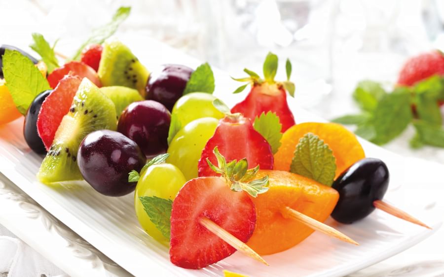 Assorted fresh fruit on skewers on a white platter: strawberries, kiwi, grapes, watermelon, orange.