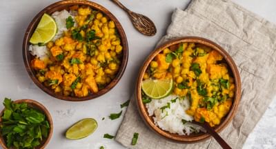Two wooden bowls of golden chickpea curry with rice, lime, and coriander leaves.