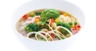 Bowl of clear vegetable noodle soup topped with a basil leaf, broccoli, peas, peppers and onions.