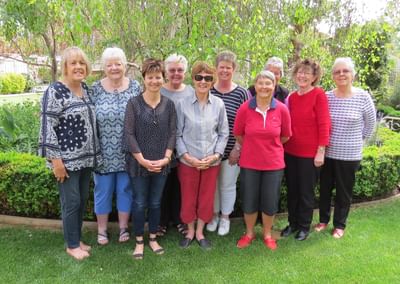 Group of adults posing in a sunny garden, standing in a row among green shrubs and trees.