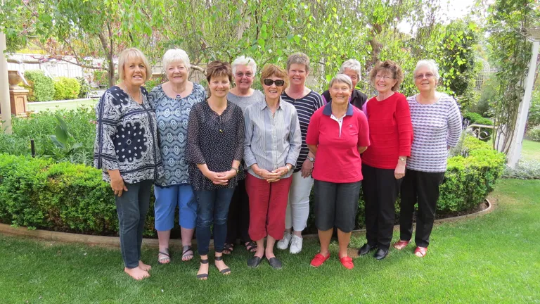 Group of adults posing in a sunny garden, standing in a row among green shrubs and trees.