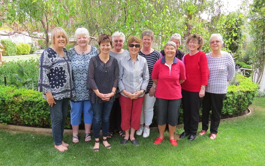 Group of adults posing in a sunny garden, standing in a row among green shrubs and trees.