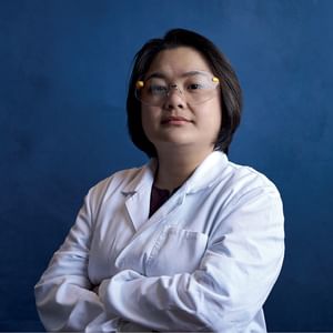 Person in a white lab coat and safety glasses stands with arms crossed against a blue backdrop.