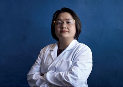 Person in a white lab coat and safety glasses stands with arms crossed against a blue backdrop.