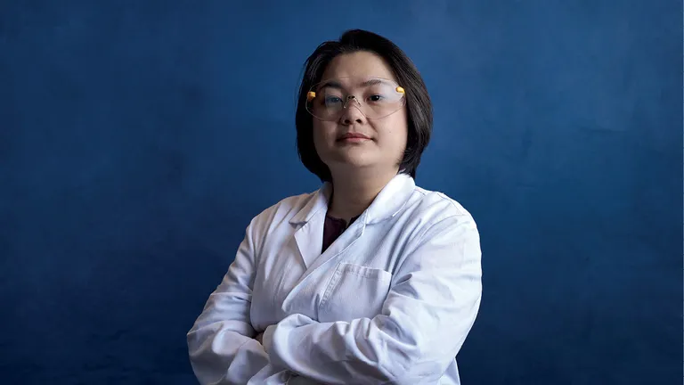 Person in a white lab coat and safety glasses stands with arms crossed against a blue backdrop.