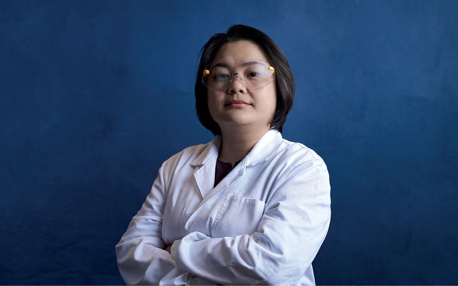 Person in a white lab coat and safety glasses stands with arms crossed against a blue backdrop.