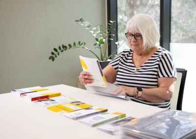 Older person with short white hair and glasses reads brochures at a white table.