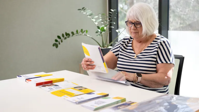 Older person with short white hair and glasses reads brochures at a white table.
