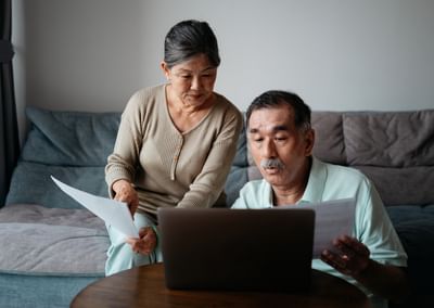 Two older adults sit on a sofa, reviewing papers while looking at a laptop.