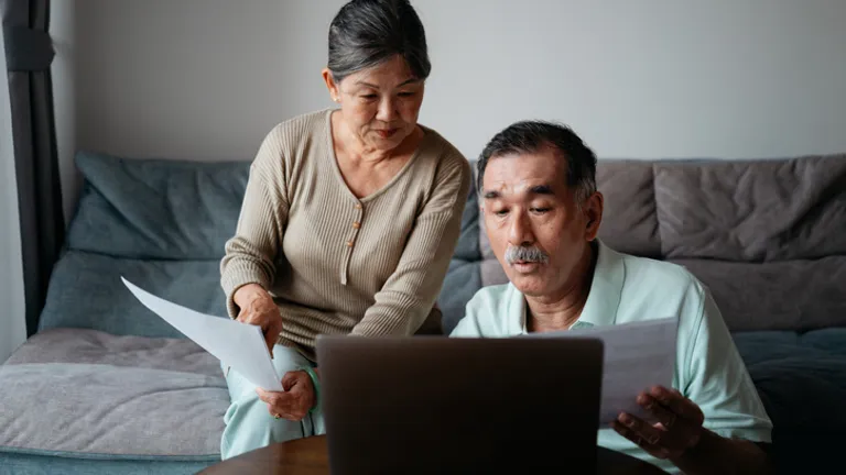 Two older adults sit on a sofa, reviewing papers while looking at a laptop.