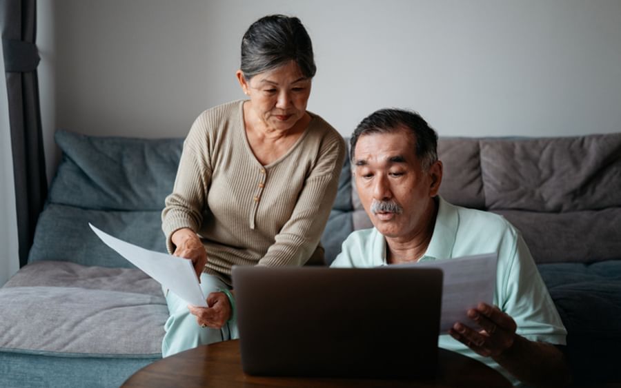 Two older adults sit on a sofa, reviewing papers while looking at a laptop.