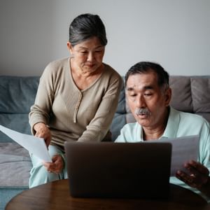 Two older adults sit on a sofa, reviewing papers while looking at a laptop.