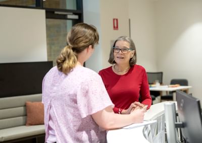 Two people chat at a bright office reception desk; one in a red top with glasses, the other in pink.