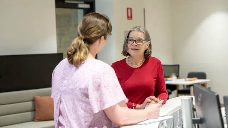 Two people chat at a bright office reception desk; one in a red top with glasses, the other in pink.