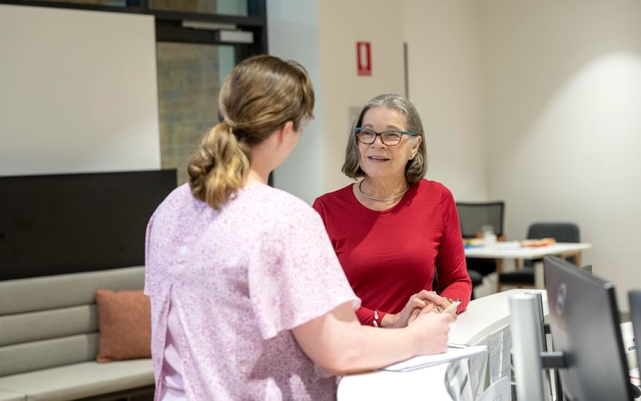 Two people chat at a bright office reception desk; one in a red top with glasses, the other in pink.