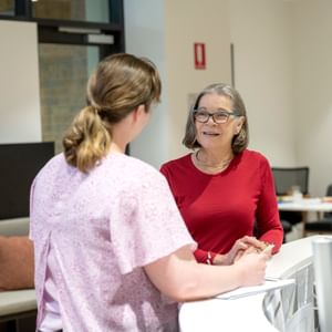 Two people chat at a bright office reception desk; one in a red top with glasses, the other in pink.