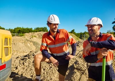 Two construction workers in orange safety vests and white helmets on a dirt site, smiling.