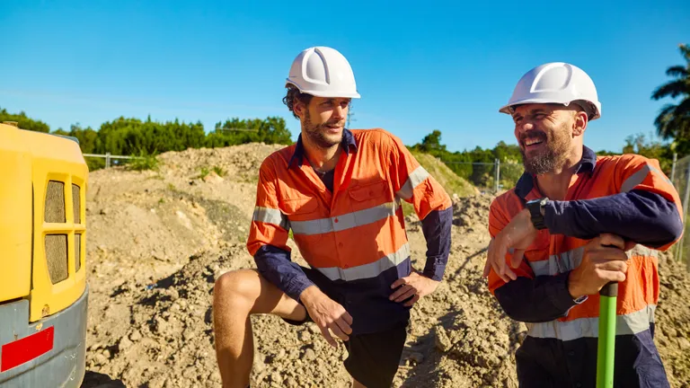 Two construction workers in orange safety vests and white helmets on a dirt site, smiling.