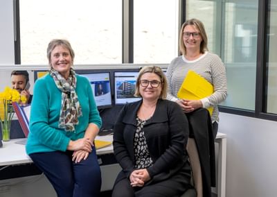 Three colleagues in an office, smiling by a desk with monitors, yellow flowers and a yellow folder.