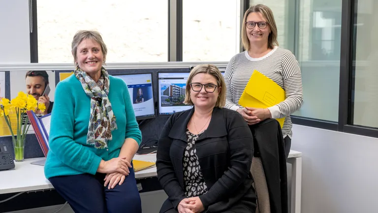 Three colleagues in an office, smiling by a desk with monitors, yellow flowers and a yellow folder.