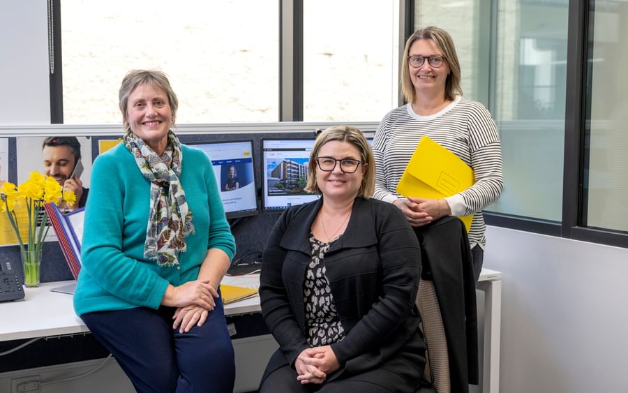 Three colleagues in an office, smiling by a desk with monitors, yellow flowers and a yellow folder.