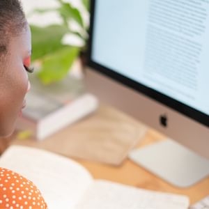 Side profile of a person in an orange polka-dot top at a desk, reading text on an iMac.