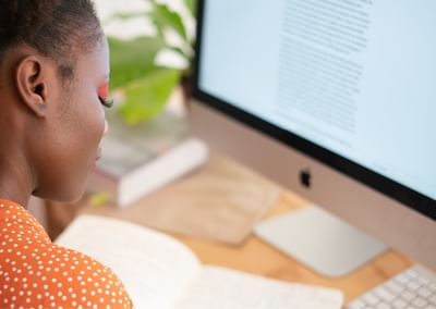 Side profile of a person in an orange polka-dot top at a desk, reading text on an iMac.