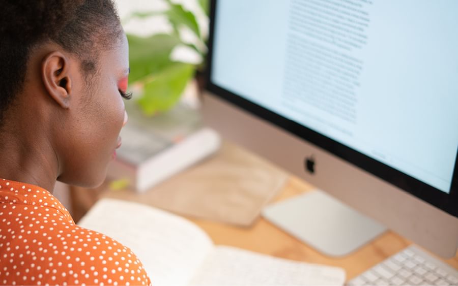Side profile of a person in an orange polka-dot top at a desk, reading text on an iMac.