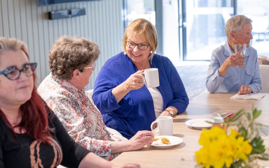 Group of seniors socialising at a light wood table with mugs, plates and yellow flowers.
