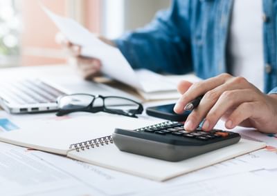 Person uses a calculator at a desk with papers, notebook, glasses and a laptop.