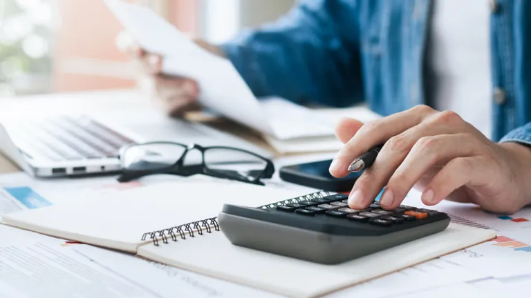 Person uses a calculator at a desk with papers, notebook, glasses and a laptop.