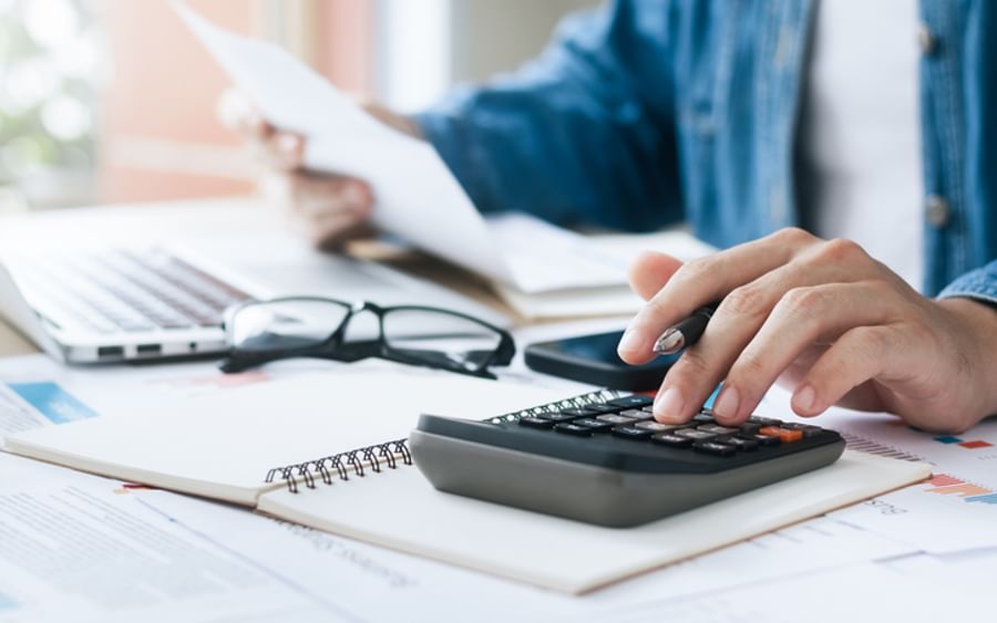 Person uses a calculator at a desk with papers, notebook, glasses and a laptop.