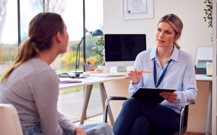 Two people in a bright office; one in a wheelchair with clipboard, the other sits opposite.