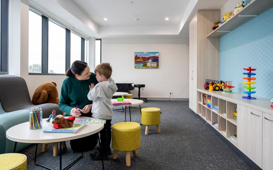 Adult and child at a round table in a bright playroom, colouring with pencils beside a teddy.