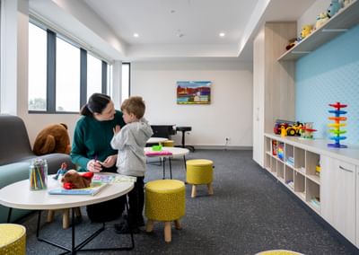 Adult and child at a round table in a bright playroom with toys, shelves, and a teddy on the sofa.