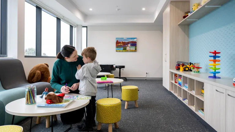 Adult and child at a round table in a bright playroom with toys, shelves, and a teddy on the sofa.