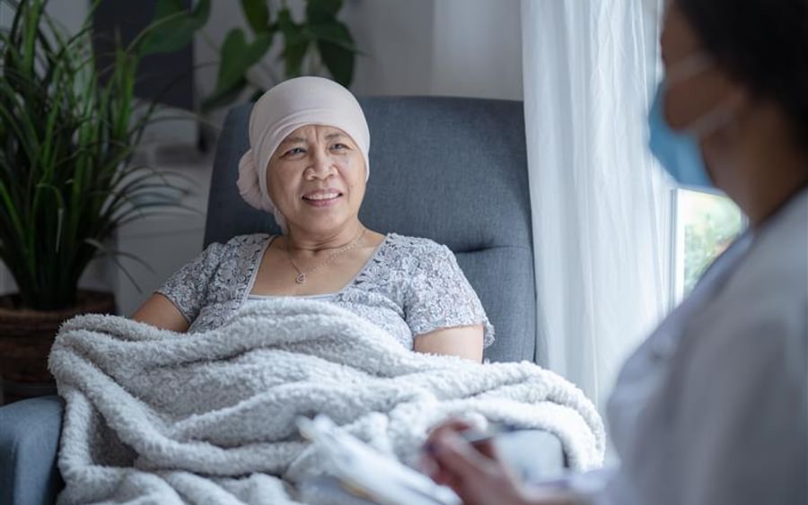 Older person sits up in a recliner with a headscarf and blanket, speaking with a masked caregiver.