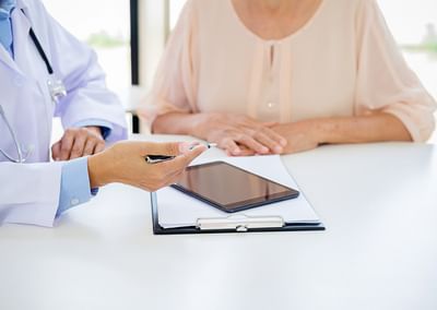 Doctor in a white coat with stethoscope consults a patient at a desk with a tablet and clipboard.