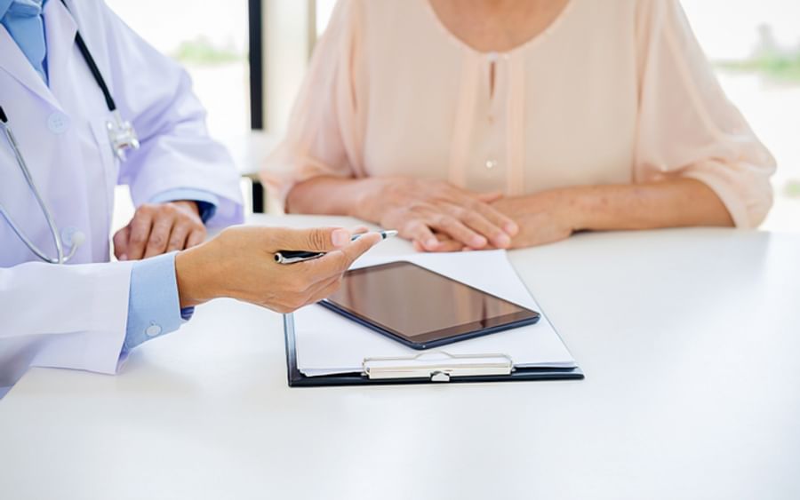 Doctor in a white coat with stethoscope consults a patient at a desk with a tablet and clipboard.