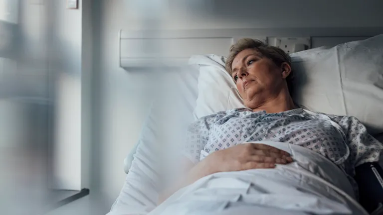 Pregnant person resting in a hospital bed, gazing out a sunlit window in a patient gown.