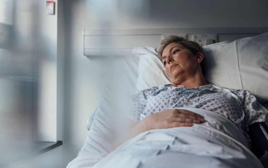 Pregnant person resting in a hospital bed, gazing out a sunlit window in a patient gown.